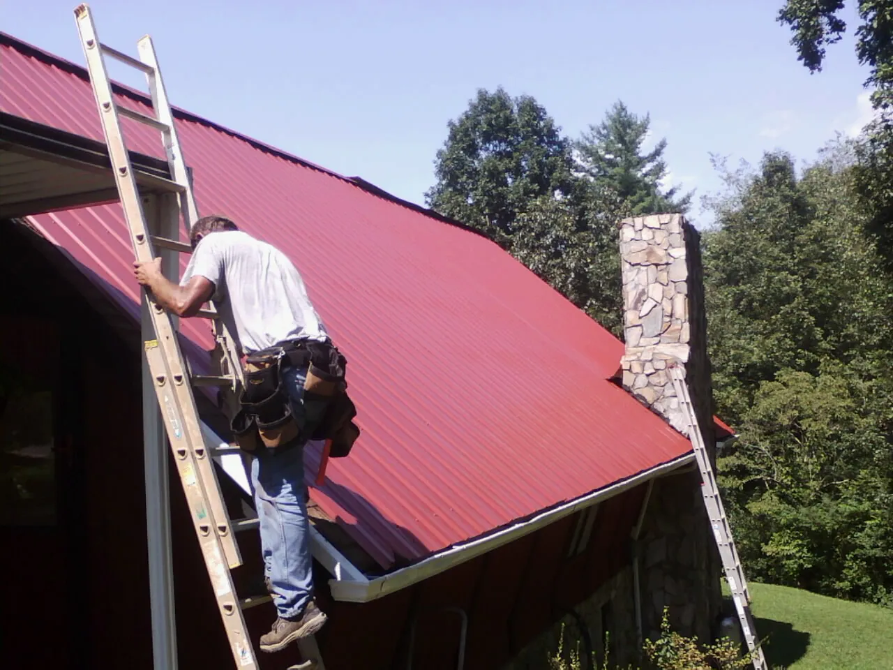 Roofer on a ladder beside a red metal roof and stone chimney — Soffit & Fascia Repair by Roofing Brothers in Hungry Horse